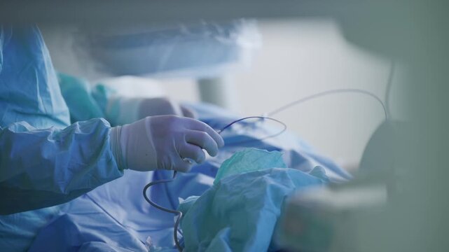 Cardiac catheterization instruments and tubing on sterile blue drape with surgeon hands preparing access for hospital surgery.