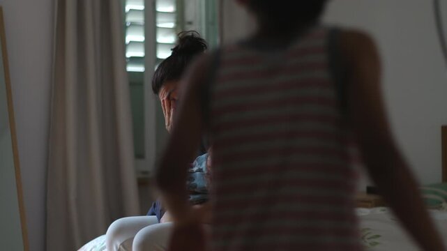 Mother sitting on bed holding toddler while showing signs of exhaustion and stress as older child seeks comfort creating emotional parenting moment of overwhelm and family pressure