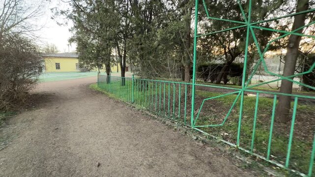 A decorative green fence borders a dirt path leading toward a yellow manor. The peaceful morning light and tall trees create a calm, nostalgic atmosphere for a stroll through the park.