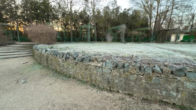 A rustic stone wall follows the curve of a garden bed covered in morning frost. Green metal arbors stand tall against the winter woods, creating a quiet and nostalgic mood in the chilly park.