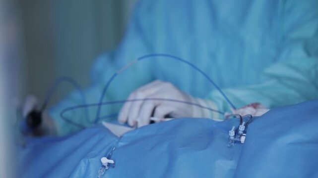Cardiac catheterization instruments and tubing on sterile blue drape with surgeon hands preparing access for hospital surgery.