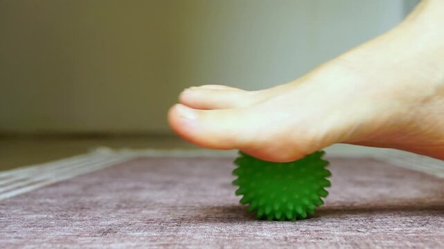 Bare female foot rolling spiky massage ball in slow circular motion in home setting. Highlights plantar pressure stimulation, trigger point activation, and self-care routine using simple tool.