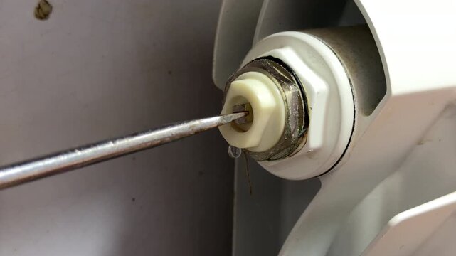 A screwdriver turns a plastic valve on a white radiator to release trapped air. A thin stream of water sprays out during this domestic maintenance task to improve heating efficiency.