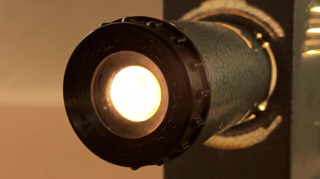 A vintage film projector whirs as it feeds a celluloid strip through its lens. The soft, warm glow from the machine creates a nostalgic and peaceful atmosphere in the dim room.