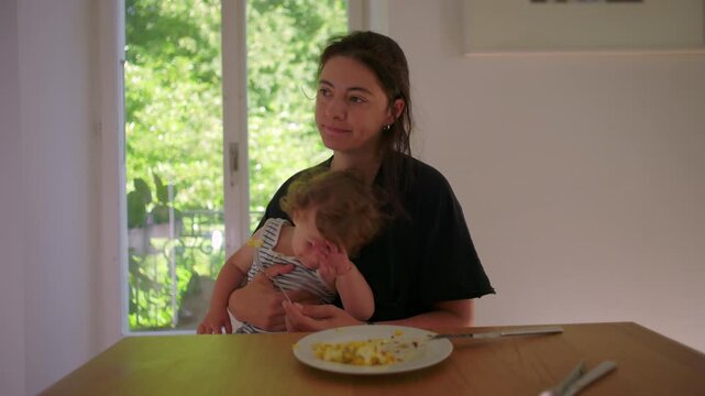 Mother trying to feed toddler at table while child turns away refusing food showing everyday parenting challenge during mealtime at home