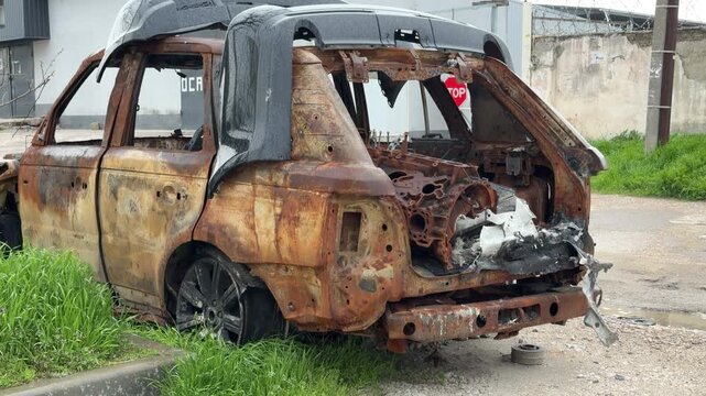 A completely gutted and charred SUV sits abandoned in the grass. The hollow metal shell and missing windows create a lonely and depressing atmosphere of urban decay.
