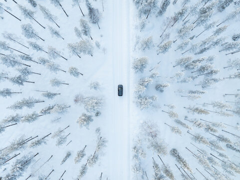 Aerial view of a dark car driving on a snowy road through a dense winter forest with frost-covered trees in Lapland, Finland.