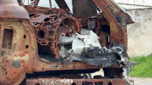 The charred and oxidized remains of a vehicle body rest in a field. This closeup shows the heavy orange rust and melted tires, creating a sad and desolate feeling of total destruction.