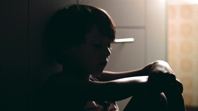 Profile view of child sitting on floor with arms resting on knees looking down expressing quiet sadness loneliness and emotional fatigue in domestic environment