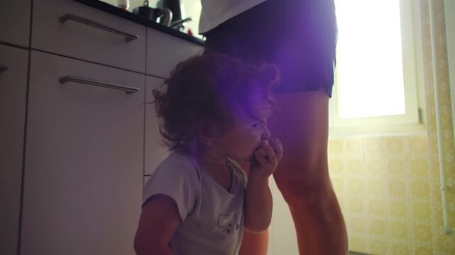 Toddler eating apple slice while standing beside mother in kitchen, natural backlit moment, everyday parenting routine, quiet domestic scene showing independence and early learning