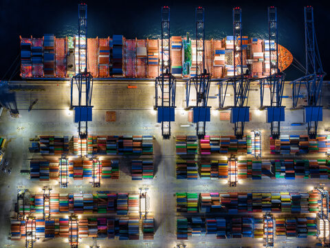 Aerial overhead view of a illuminated container terminal dock with loading and unloading activity by cranes and trucks off cargo ships during evening