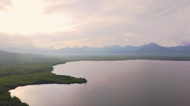 Aerial view over Iwahig in Palawan reveals a vast mangrove forest meeting a calm coastal bay. The mountain ridge of mainland Palawan rises in the background under a bright sunset sky. Warm light sprea