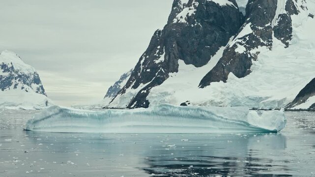 Flat tabular iceberg with perfect water reflection floating in calm Lemaire Channel Antarctica beside dramatic dark rocky cliffs