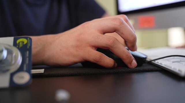 Close-up of a hand operating a computer mouse on an office desk beside a keyboard, with a blurred monitor in the background and soft indoor lighting.