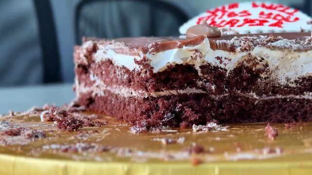 Close-up of a partially eaten chocolate cream cake slice on a golden cake board, showing soft layers, frosting texture, crumbs, and red icing detail in natural light.