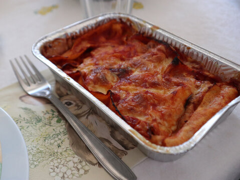 Porzione di parmigiana di melanzane o lasagna al forno calda servita in una vaschetta monouso di alluminio da asporto posizionata sulla tavola