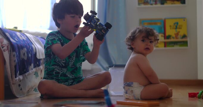 Two children sitting on floor playing with toy vehicle and blocks, older child focused on building while baby looks toward camera, interaction showing sibling bonding and playful learning
