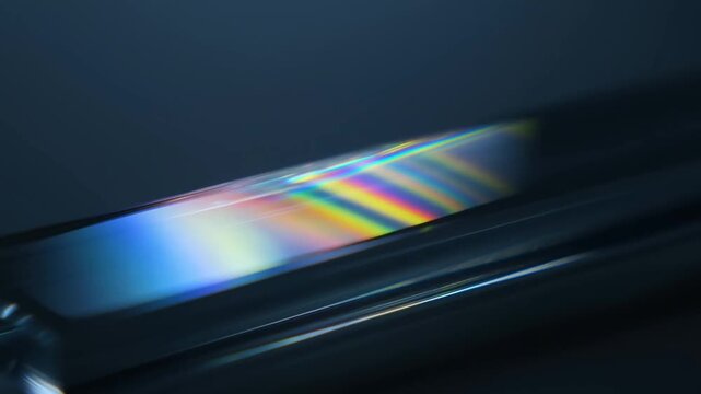 Slow tracking shot of angled glass prism reflecting rainbow light across glossy surface on dark studio tabletop with subtle iridescent highlights