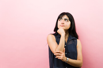 Latin woman contemplating on pink background, thinking seriously, making a decision, looking up for...