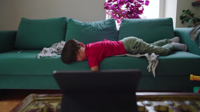 Young boy lying on sofa watching tablet screen, tired posture, absorbed attention, relaxed home environment, quiet moment of screen time and digital entertainment in living room