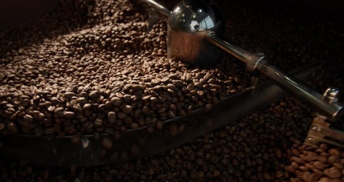 Slow motion roasted coffee beans tumbling inside industrial roaster drum with rotating metal arm.