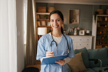 A home health nurse stands in a living room smiling while holding a clipboard. She is dressed in blue scrubs and provides care to her patient in a comfortable setting. © Stockphotodirectors