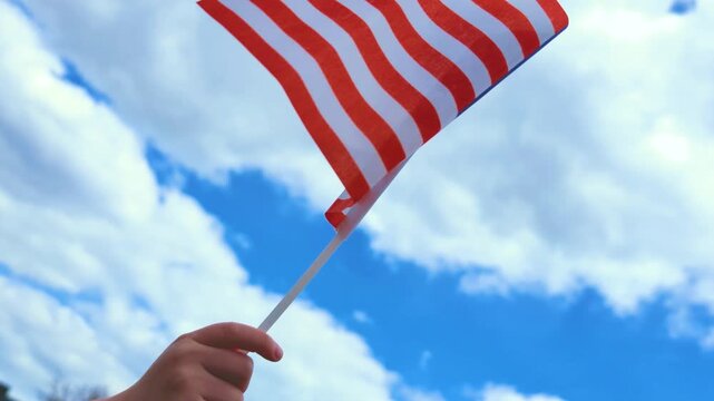 Video of child hand holding and waving USA flag against blue sky with clouds. Patriotic outdoor scene perfect for Independence Day, celebrations and American themed projects.