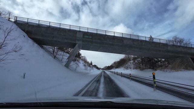 Point of view driving on a snowy highway during light snowfall, moving through an underpass with forest surroundings and cloudy sky. Smooth transition scene with no traffic
