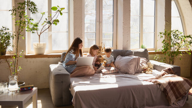 Wide shot of mother and two children using laptop and tablet on sofa in bright living room. Concept of hybrid learning, multi device usage, home routine, technology integration and modern family life.