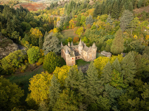 Aerial view of Chateau du Champ, a historic stone castle with pointed towers nestled among vibrant green and yellow autumn trees in a lush valley Altier, Occitanie, France.