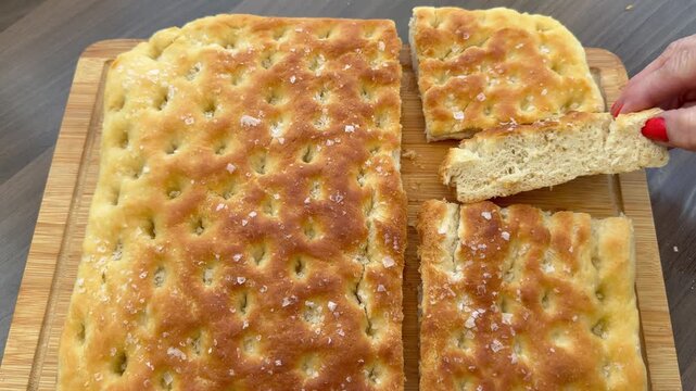 Closeup of a woman&rsquo;s hand taking a cut piece from a batch of oven baked, focaccia Italian bread, a classic recipe of flat, dimpled dough topped with rosemary, olive oil and sea salt flakes.