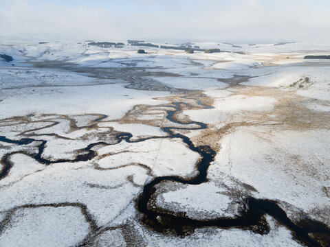 Aerial view of a winding river meander through snow-covered fields and rolling hills in the Aubrac plateau near Nasbinals, Occitanie, France.