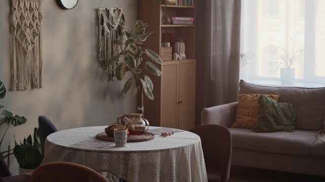 Medium long shot of boho living room interior with macrame decor on wall, table with mugs and teapot, sofa, chairs and green plants, no people