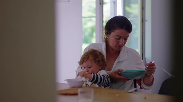 Mother eating meal while toddler plays with food at kitchen table, candid parenting moment showing multitasking, patience, and everyday family routine with emotional connection