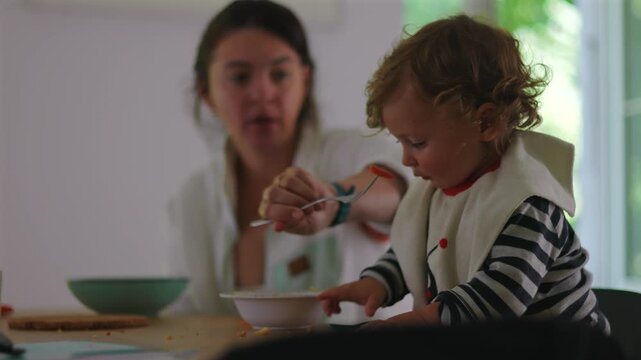 Mother feeding toddler with spoon while child simultaneously eats with hand, dynamic mealtime moment showing mixed independence, curiosity, parenting during everyday routine