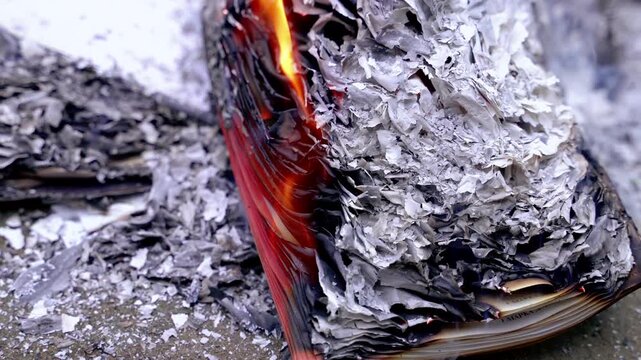 Open book catching fire on a stone surface, surrounded by heaps of gray paper ash and embers. Open book burning on the ground with gray ash.