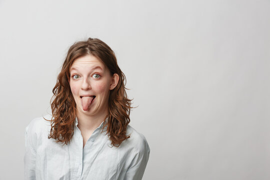 Young woman with curly hair wearing white shirt sticks out tongue playfully, isolated on light background. Concept of fun, humor, and casual lifestyle portrait.