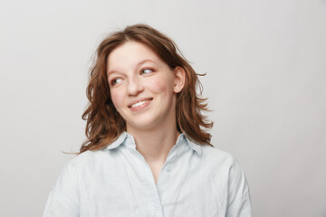 Fototapeta premium Smiling young woman with wavy brown hair wearing a white shirt looks sideways on a light background. Concept of happiness, casual style, and natural beauty portrait.