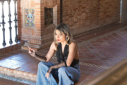 A young, attractive woman wearing jeans, gloves and a black T-shirt, smoking a cigarette whilst sitting on the steps of Spain Square in Seville. The woman is on holiday in the city.