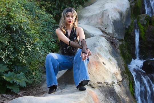 A young, pretty woman, wearing make-up and dressed in jeans, gloves and a black T-shirt, sitting on the rocks beside a crystal-clear waterfall surrounded by lush vegetation.