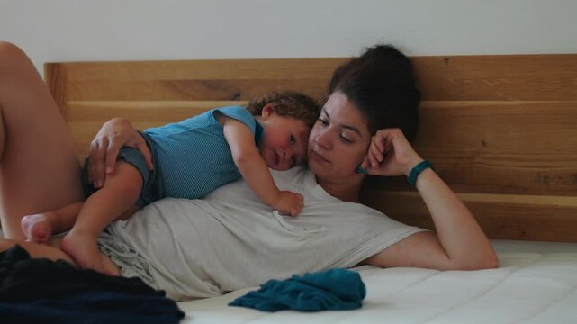 Mother lying on bed with toddler resting on her chest expressing exhaustion emotional fatigue and quiet weight of caregiving while maintaining deep maternal bond in intimate home setting