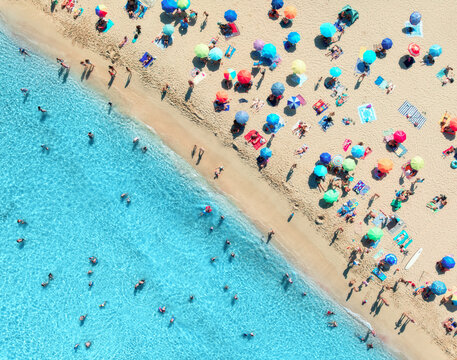 Aerial view of colorful umbrellas on crowded sandy beach, swimmers in blue sea, green trees on sunny summer day. Mallorca, Balearic Islands, Spain. Tropical top drone view. Turquoise water. Travel