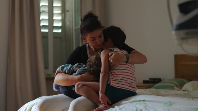 Mother sitting on bed embracing two children close together offering comfort during difficult moment showing warmth care emotional support and quiet resilience in family life