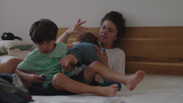 Mother sitting on bed holding toddler while older child rests beside her expressing fatigue emotional weight and quiet challenge of daily motherhood within intimate family home setting