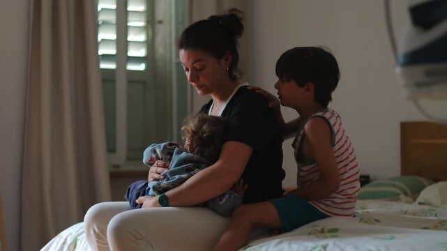 Mother sitting on bed holding toddler with tired expression while older child cries and leans on her showing emotional overwhelm parenting stress and demanding family moment