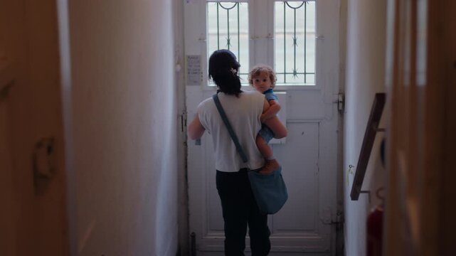 Mother walking down narrow hallway carrying toddler toward front door preparing to step outside capturing everyday routine of leaving home with child and quiet sense of care responsibility