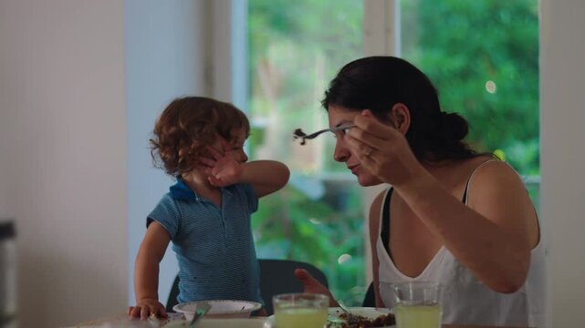 Mother trying to feed toddler during mealtime while child resists and turns away expressing independence frustration and early autonomy in everyday parenting interaction at home