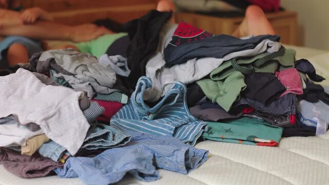 Pile of folded and unfolded clothes on bed while family rests in background showing everyday domestic life routine and household tasks within lived in home environment