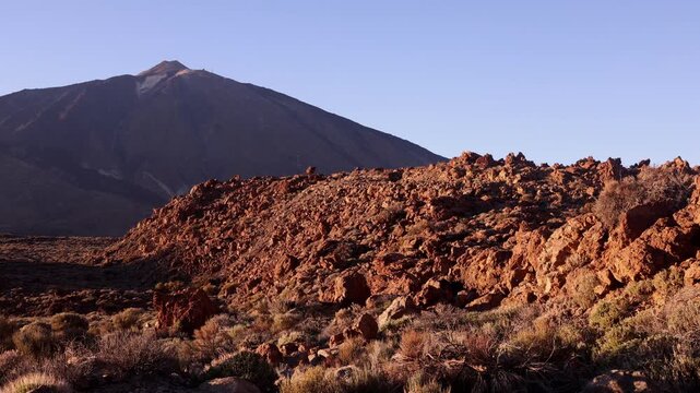 Panoramic view of Mount Teide rising above dramatic red lava rock field in Tenerife. Warm sunset light on volcanic terrain under clear blue sky &mdash; iconic Canary Islands travel scene.