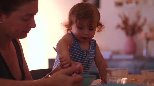 Toddler eating with fork at table while mother sits nearby guiding and observing showing independence learning and supportive parenting during everyday family mealtime at home